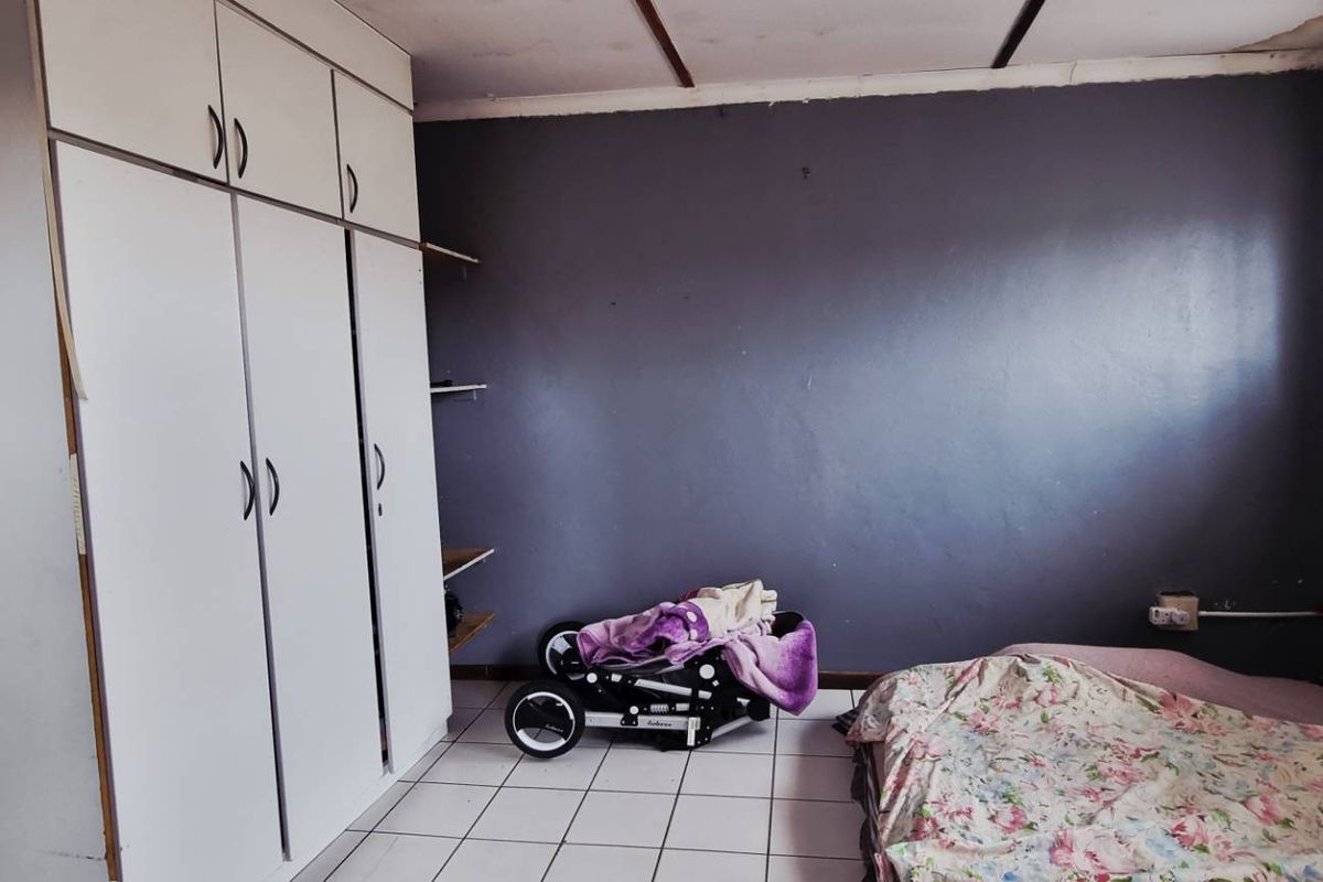 A bedroom featuring built-in cupboards, tiled flooring, and a window with security bars. The room requires cosmetic updates to the ceiling and walls.