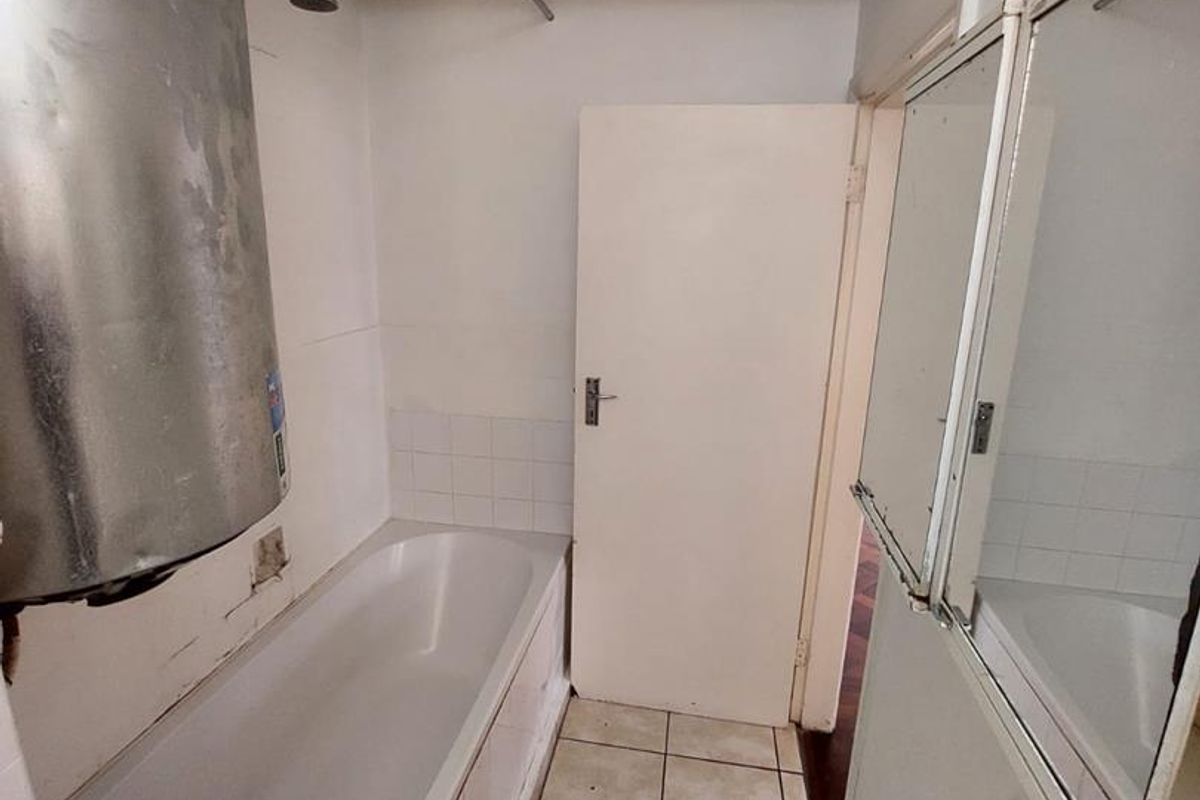 A compact bathroom featuring a bathtub, wall-mounted shower head, and an exposed electric water heater, with tiled flooring and wall-mounted mirrors.