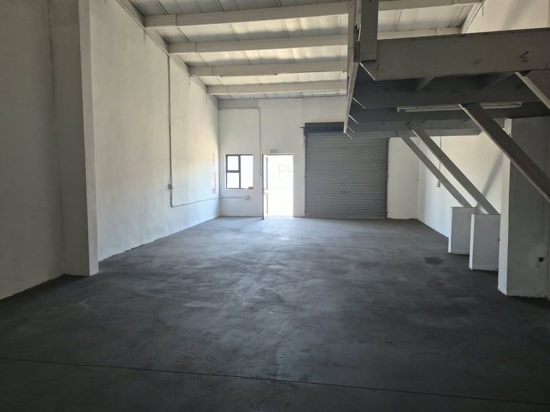 Spacious industrial warehouse unit featuring high ceilings with exposed trusses, a roller shutter door, and a functional mezzanine level for additional storage or office space.