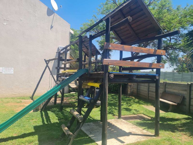 A communal garden area featuring a wooden jungle gym with a slide and a fixed bench, set against a concrete boundary wall.