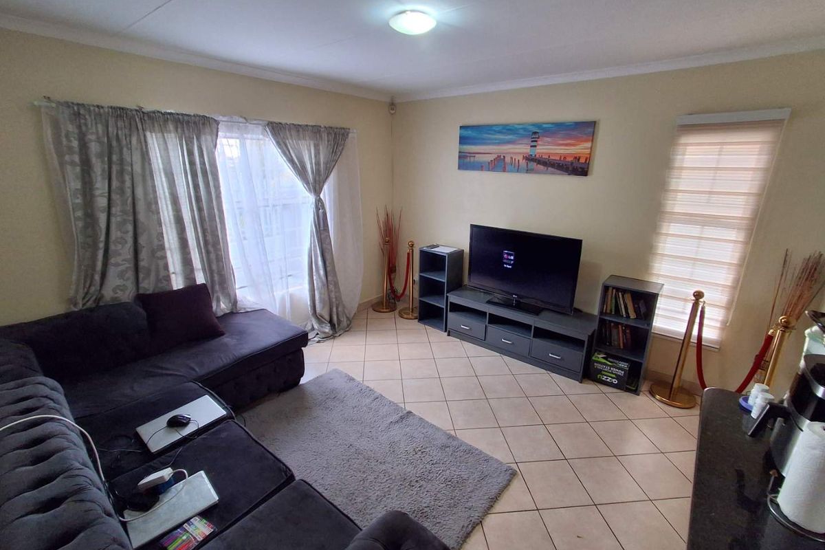 A living room featuring tiled flooring, neutral wall finishes, and window treatments including roller blinds and curtain tracks.