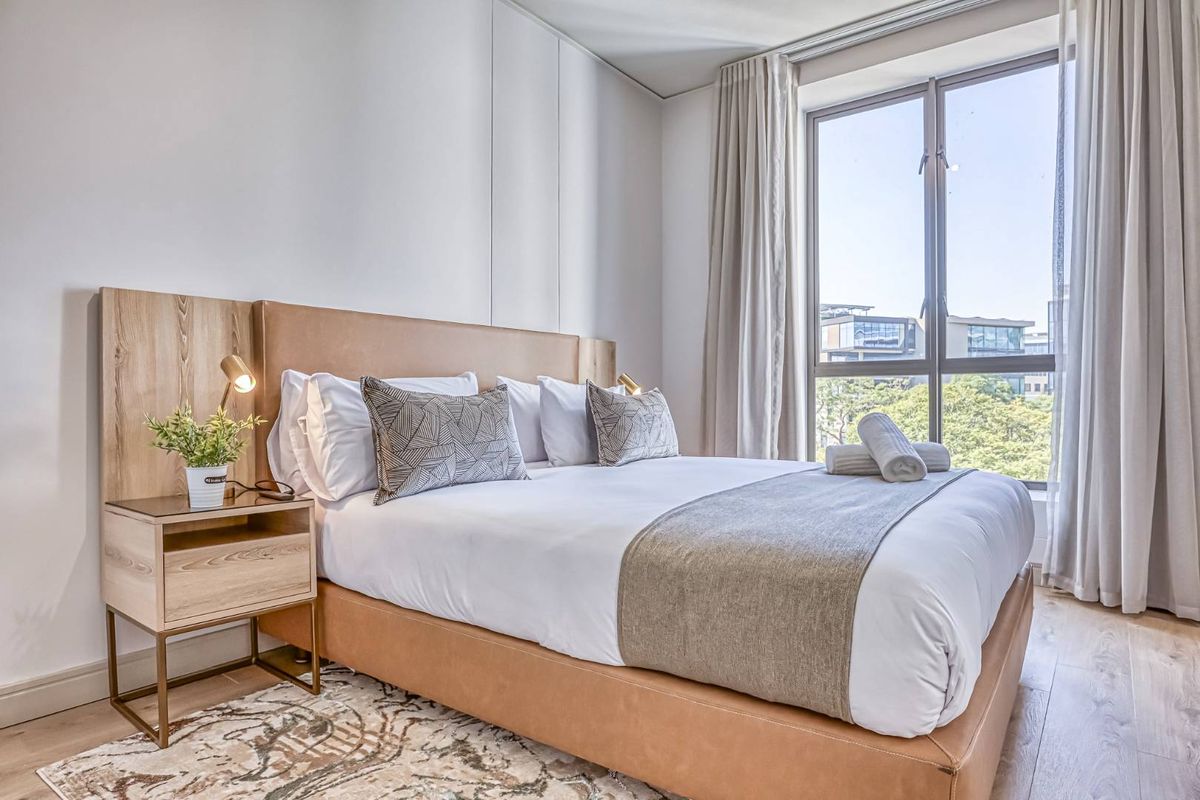 A bright and modern bedroom featuring light wood-look flooring, neutral walls, and a large window providing natural light and urban views.