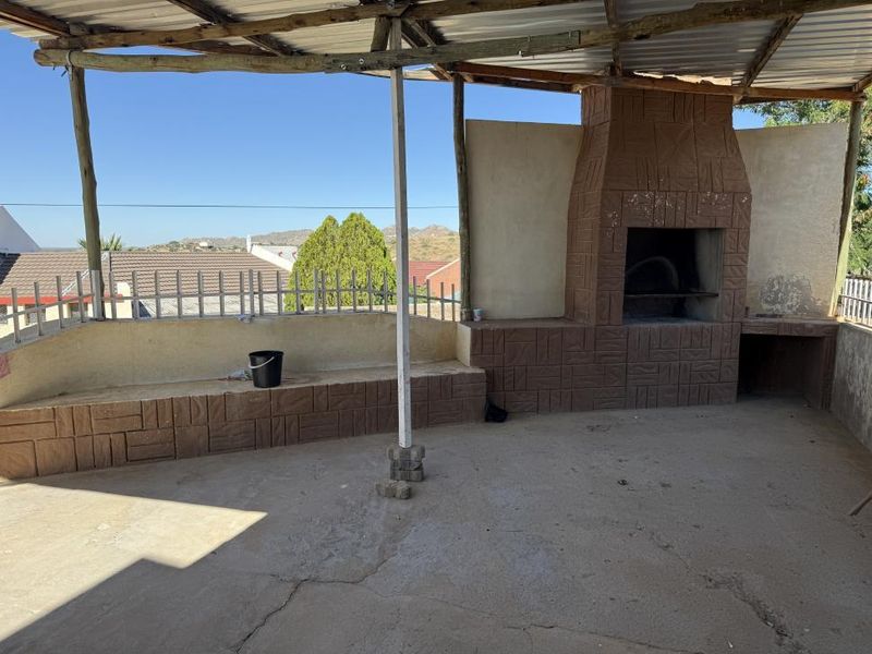 Covered outdoor patio area featuring a built-in braai, concrete flooring, and a rustic timber-supported roof structure with views of the surrounding neighborhood.