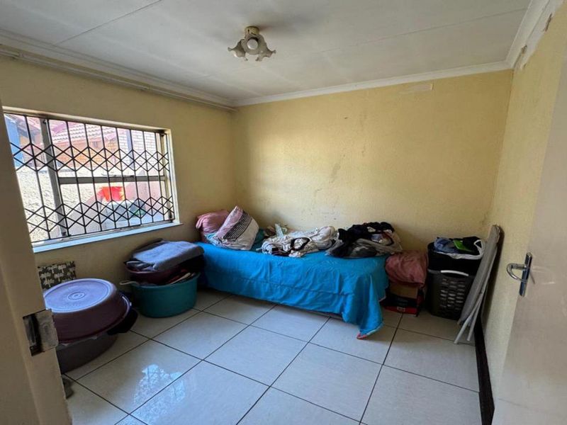 A compact bedroom featuring tiled flooring, a security-gated window, and basic ceiling lighting, requiring cosmetic updates to improve its overall appeal.
