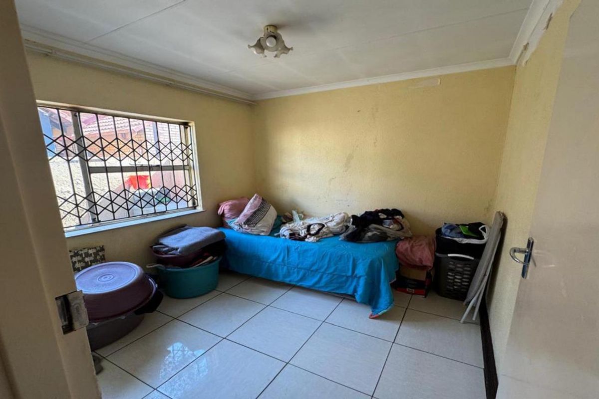A compact bedroom featuring tiled flooring, a security-gated window, and basic ceiling lighting, requiring cosmetic updates to improve its overall appeal.