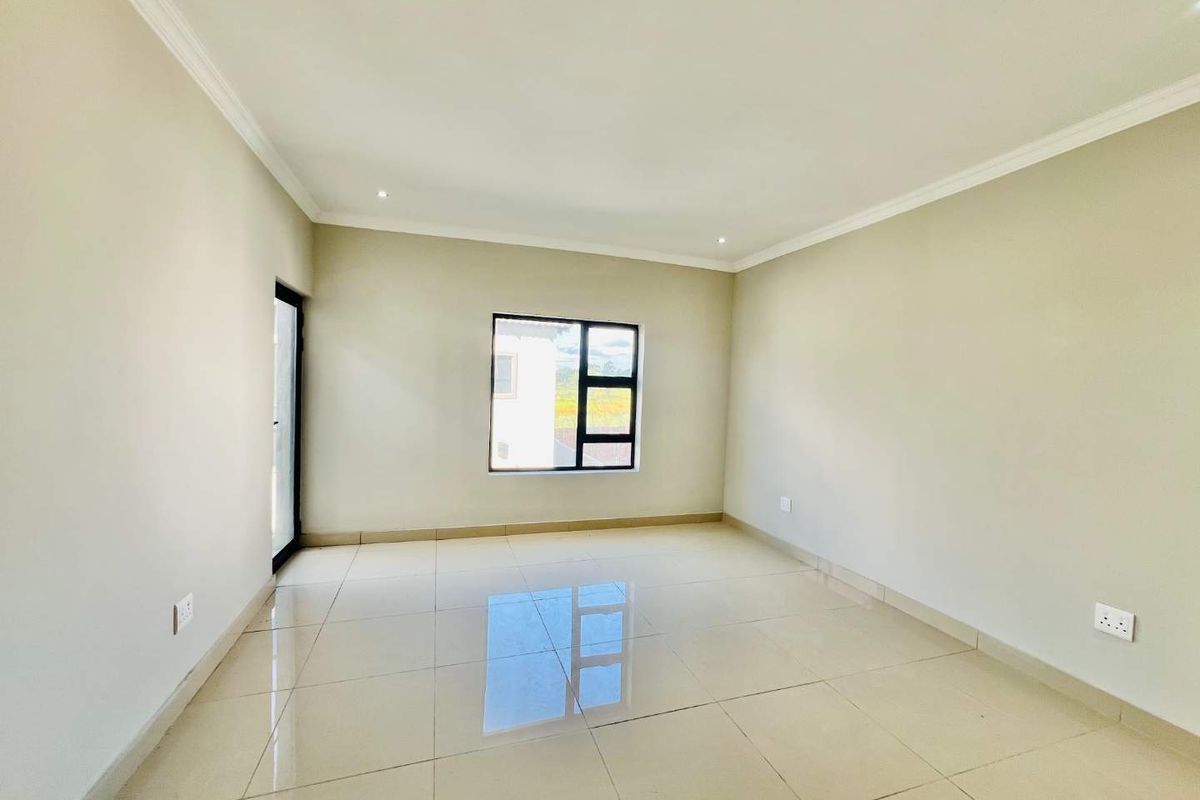 A bright, empty living room featuring neutral tiled flooring, recessed ceiling lighting, and a large window providing natural light.
