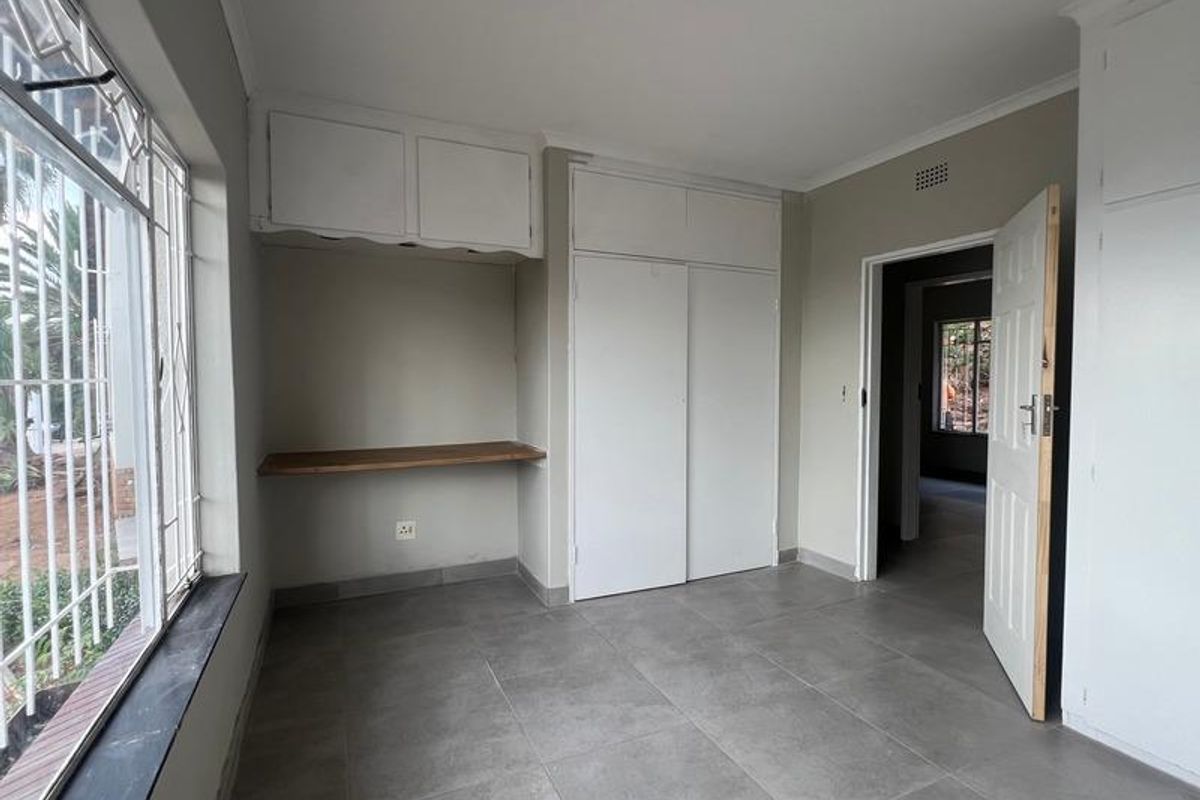 A bedroom featuring built-in cupboards, a dedicated desk area, tiled flooring, and large windows with security bars.