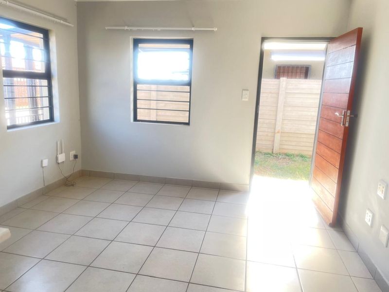 A bright, empty bedroom featuring tiled flooring, aluminium window frames with security bars, and a timber door leading to the exterior.