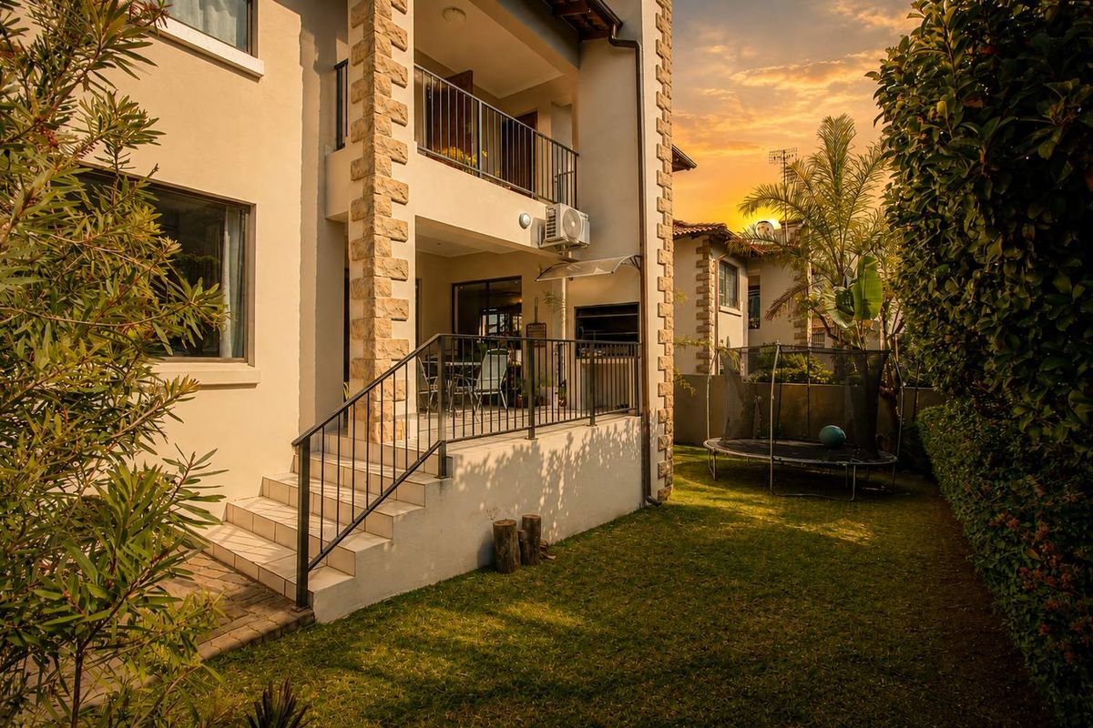 Rear view of the townhouse featuring a covered patio with built-in braai, stone-clad pillars, metal railings, and a private garden area.
