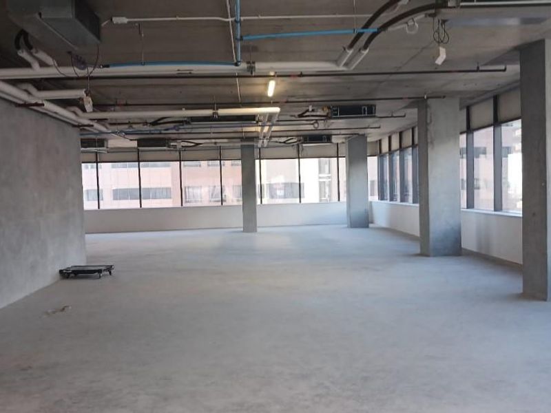 A spacious, open-plan commercial shell featuring exposed concrete ceilings, industrial piping, concrete columns, and expansive windows providing ample natural light.