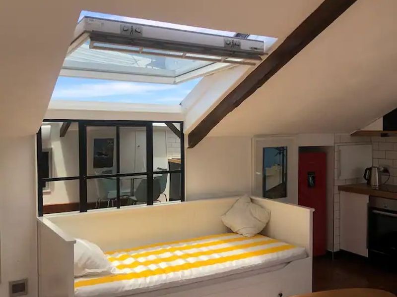 A bright open-plan attic apartment featuring exposed timber beams, a large skylight, timber flooring, and a modern kitchen with subway tile backsplash.