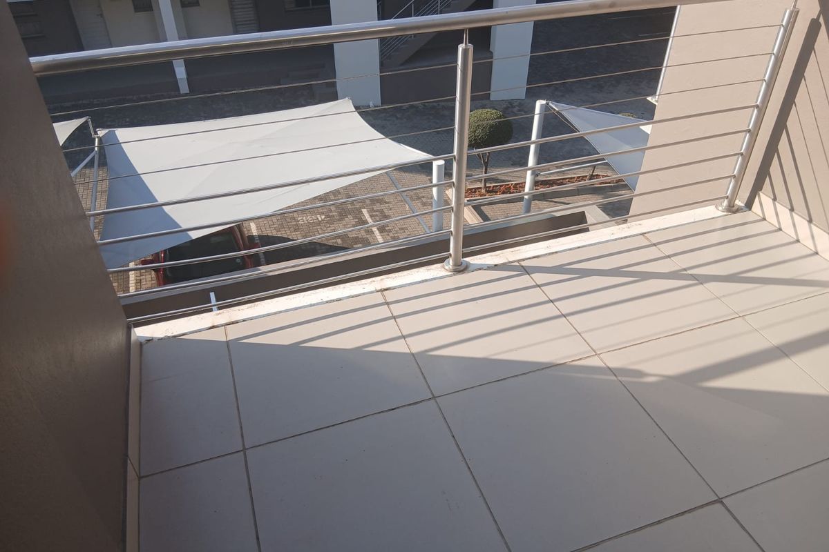 A clean, tiled balcony featuring a modern stainless steel balustrade, overlooking the apartment complex parking area with shade sails.