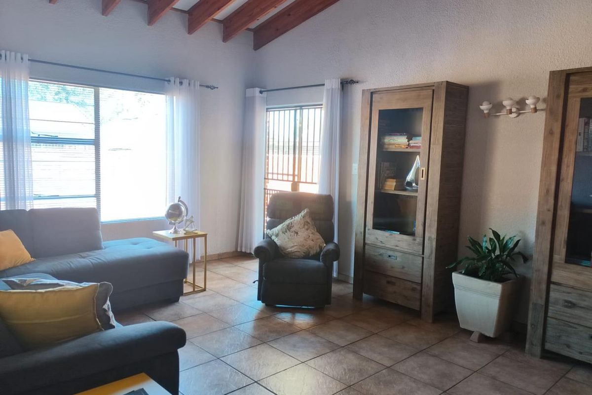 A living room featuring exposed timber ceiling beams, ceramic floor tiles, and natural light through large windows and a sliding door with security gate.