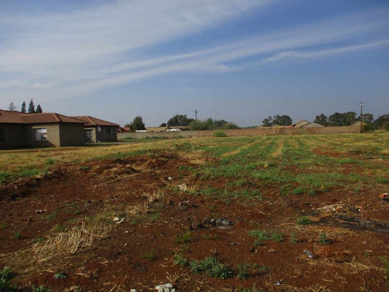 A vacant residential plot of land featuring open ground and a boundary wall, situated in a developed residential neighborhood.