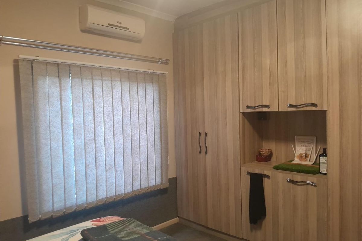 A bedroom featuring light wood-grain built-in cupboards, a wall-mounted air conditioning unit, and vertical window blinds.