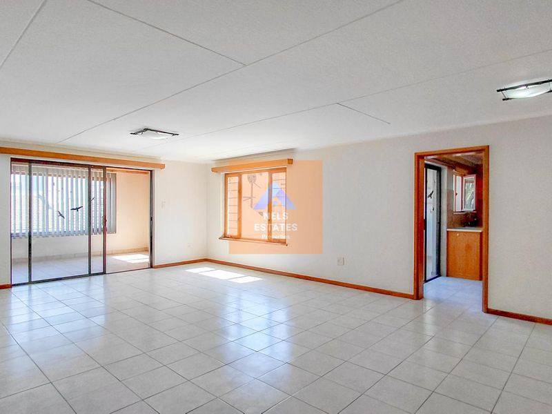 Spacious, empty living area featuring neutral tiled flooring, wooden window frames, and access to an adjoining balcony through sliding glass doors.
