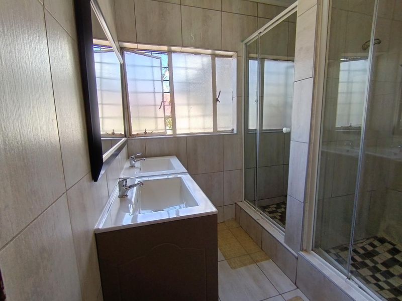 A functional bathroom featuring a double vanity unit, a glass-enclosed shower, and neutral wall and floor tiling.