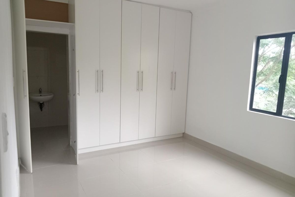 A bright, empty bedroom featuring modern white built-in cupboards, neutral tiled flooring, and an adjoining en-suite bathroom.