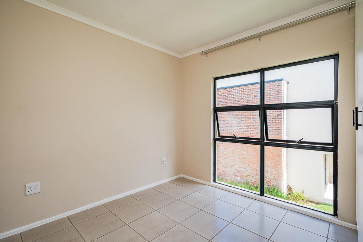 A bright, empty bedroom featuring neutral tiled flooring, beige walls, and a large black-framed window overlooking an exterior brick wall.
