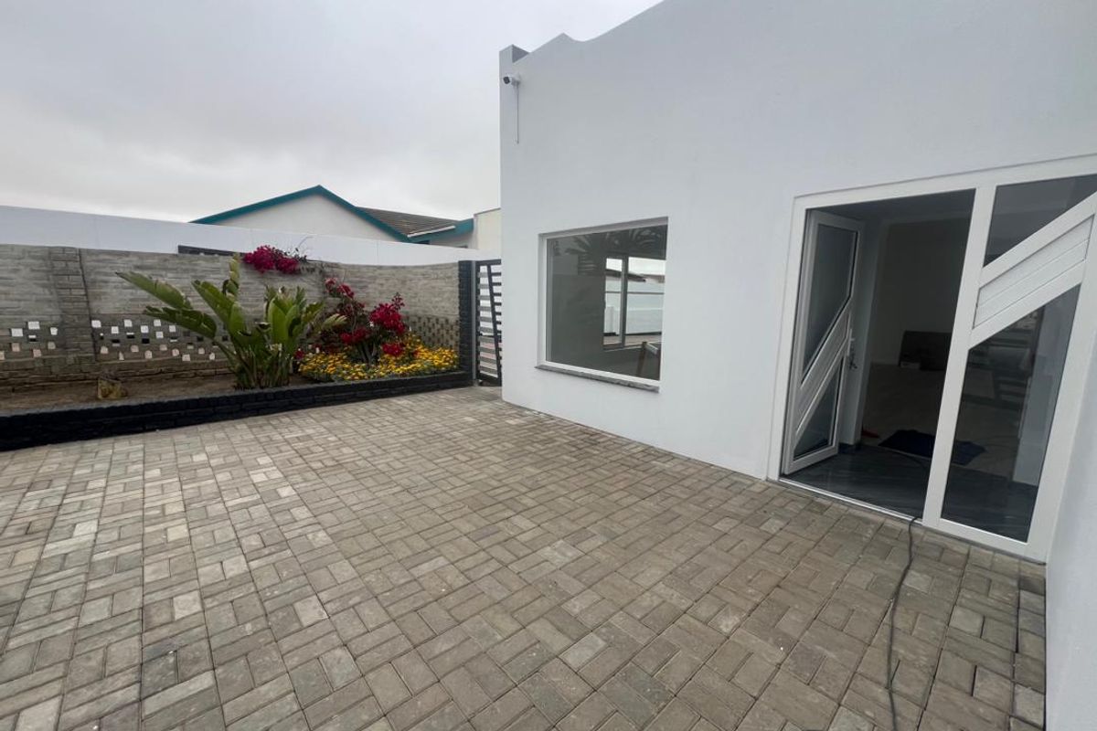 A clean, paved rear patio area featuring a white exterior wall, modern glass door access, and a landscaped garden bed with a decorative boundary wall.