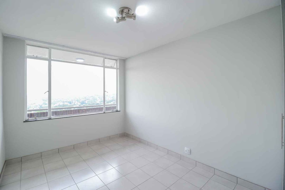 A bright, empty bedroom featuring neutral tiled flooring, a large window with natural light, and a built-in wardrobe.