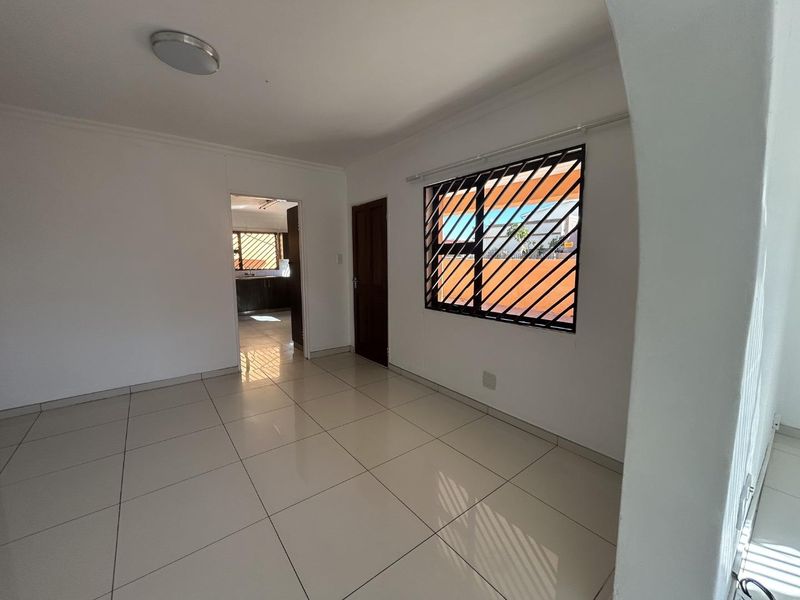 A bright, empty living area featuring neutral tiled flooring, an arched entryway, and security bars on the window, leading through to the kitchen.