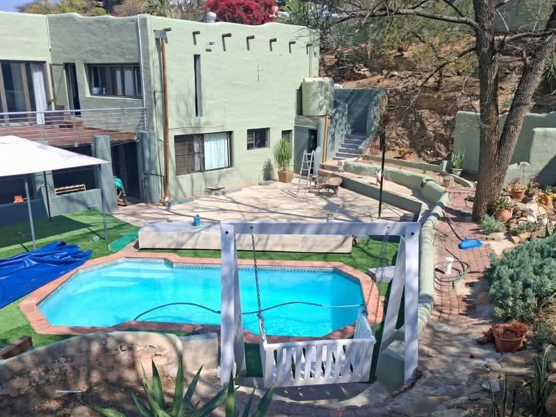 Rear exterior view featuring a swimming pool, paved patio area, elevated balcony, and tiered garden landscaping with retaining walls.