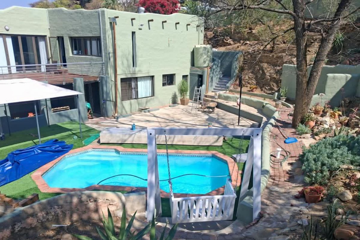 Rear exterior view featuring a swimming pool, paved patio area, elevated balcony, and tiered garden landscaping with retaining walls.
