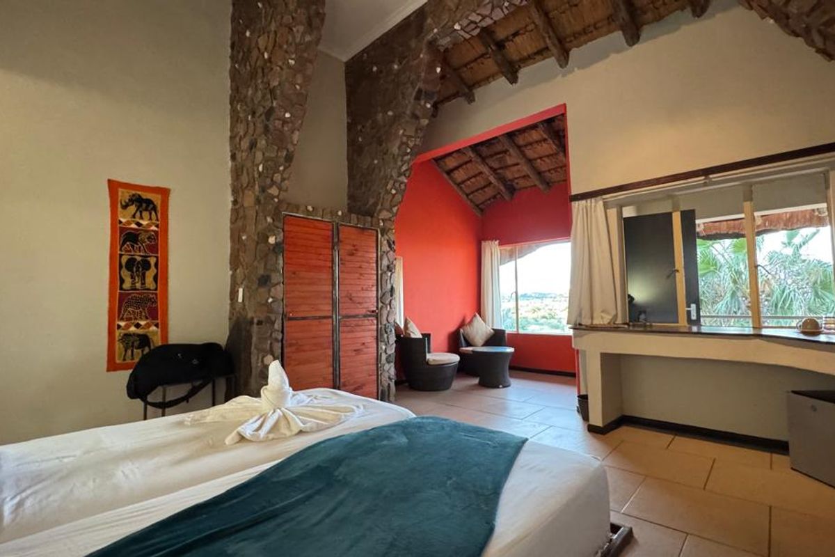 Spacious lodge bedroom featuring rustic stone archways, high thatched ceilings, tiled flooring, and an integrated desk area with views.