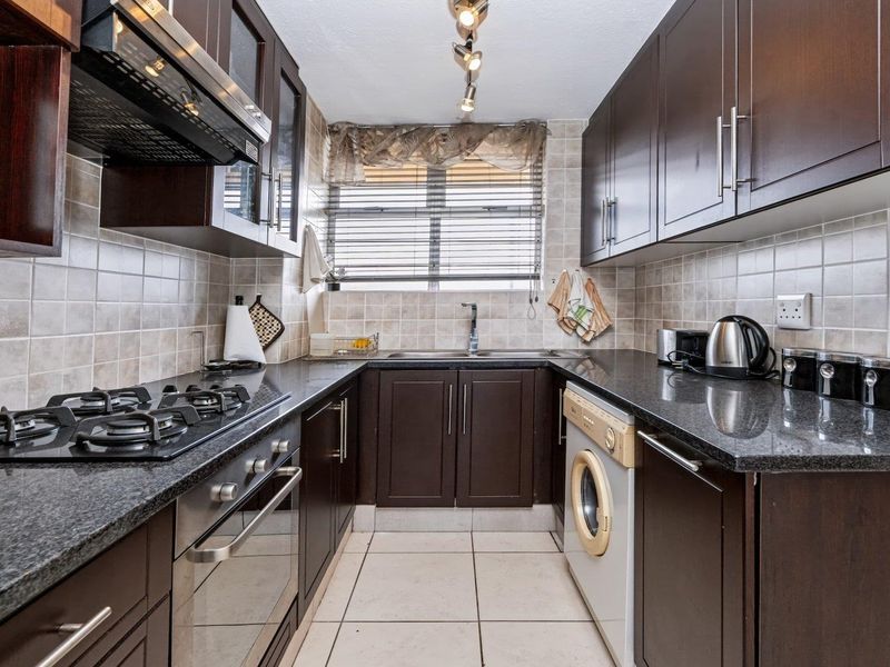 Compact kitchen featuring dark wood cabinetry, granite countertops, gas hob, built-in oven, and integrated washing machine with neutral tiled finishes.