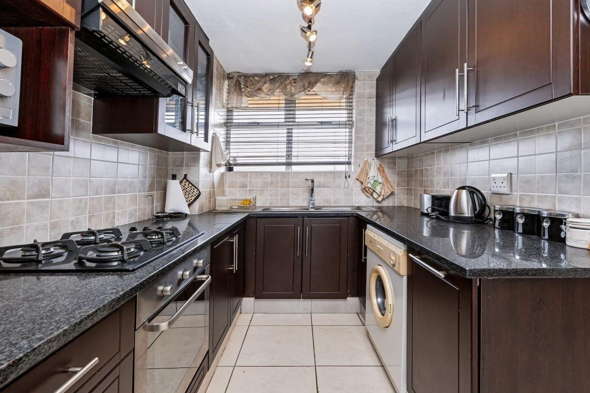 Compact kitchen featuring dark wood cabinetry, granite countertops, gas hob, built-in oven, and integrated washing machine with neutral tiled finishes.