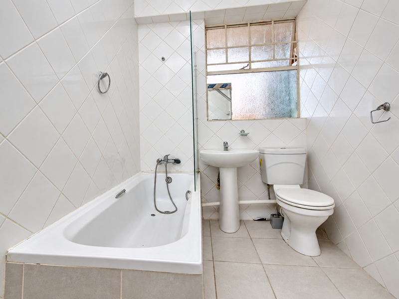 A compact bathroom featuring a bathtub with a glass screen, pedestal basin, and toilet, finished with white wall tiles and neutral floor tiles.