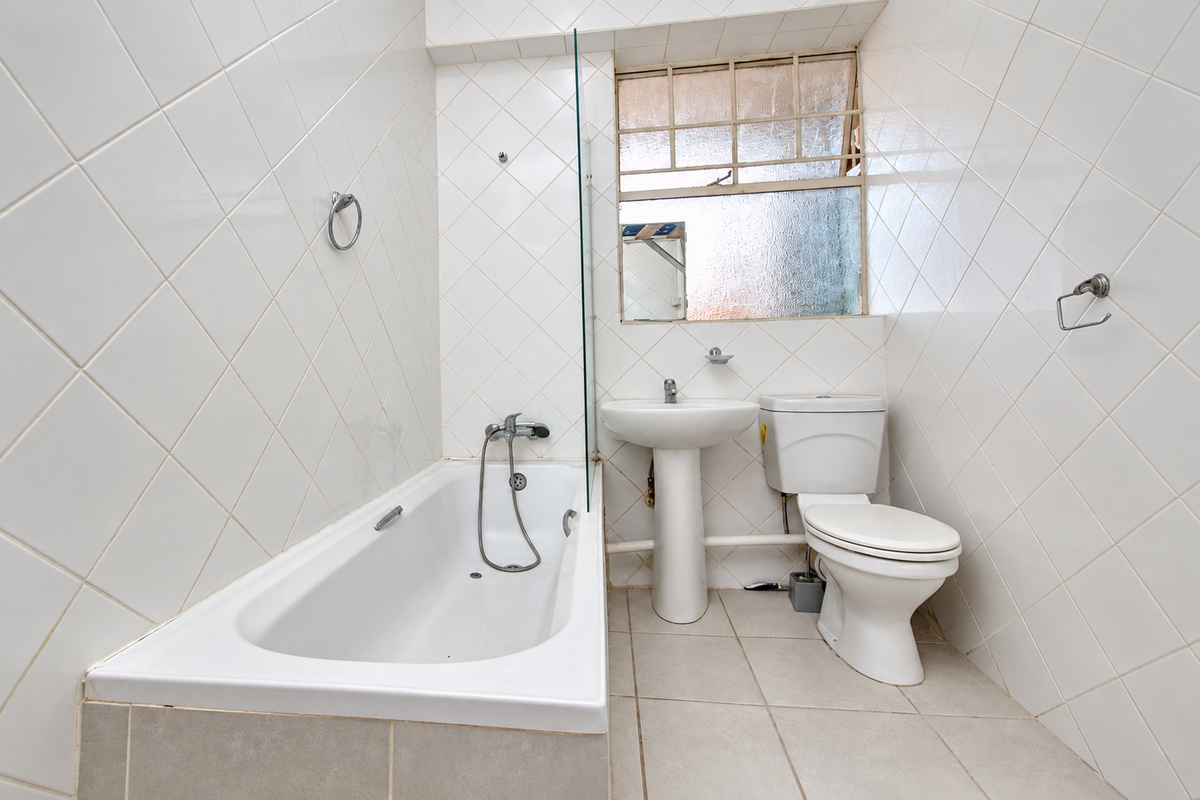 A compact bathroom featuring a bathtub with a glass screen, pedestal basin, and toilet, finished with white wall tiles and neutral floor tiles.