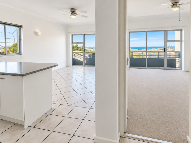 Bright open-plan living area featuring tiled kitchen space, carpeted lounge, and sliding doors leading to a balcony with ocean views.