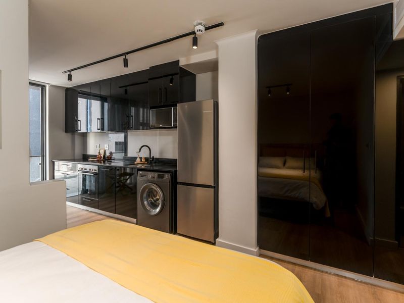 Modern open-plan studio apartment featuring sleek black high-gloss kitchen cabinetry, integrated appliances, track lighting, and floor-to-ceiling mirrored built-in wardrobes.