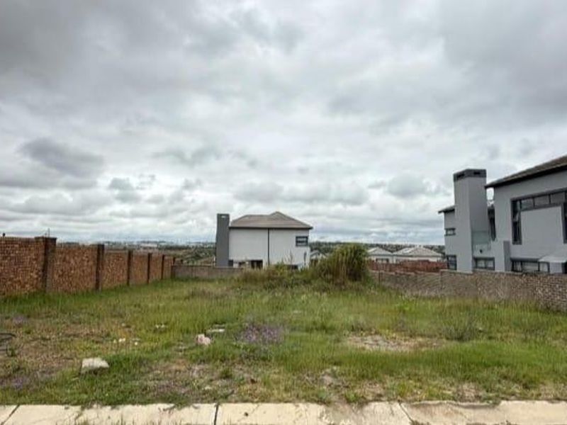 A vacant residential plot featuring a brick boundary wall, situated in a developed neighborhood with surrounding modern homes.