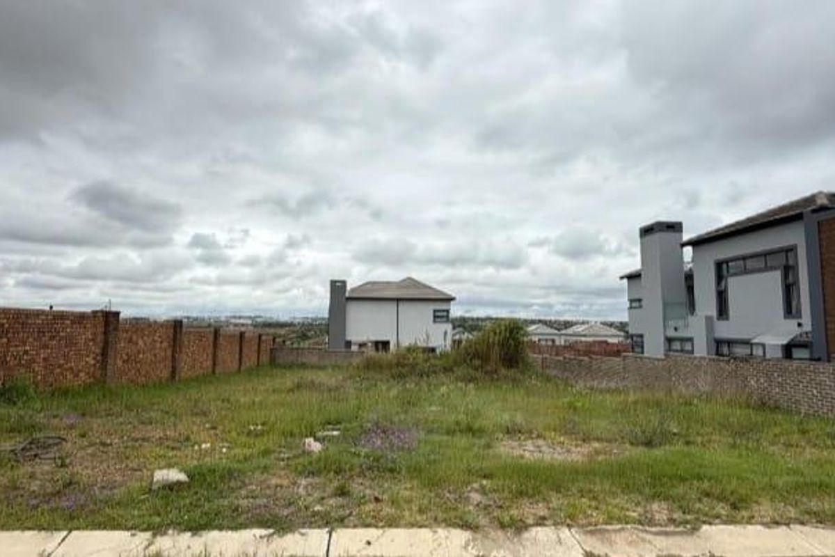 A vacant residential plot featuring a brick boundary wall, situated in a developed neighborhood with surrounding modern homes.