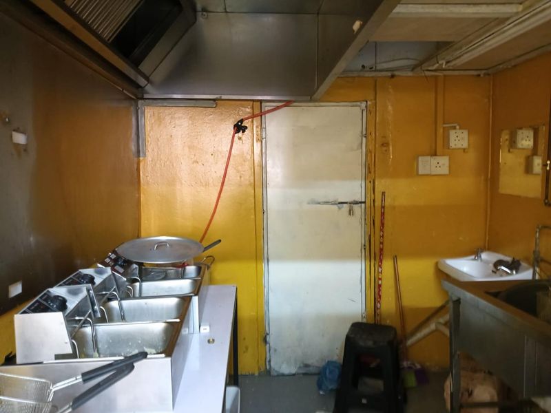 A commercial kitchen space featuring stainless steel deep fryers, an extractor hood, and a sink, with dated yellow wall finishes and exposed electrical infrastructure.