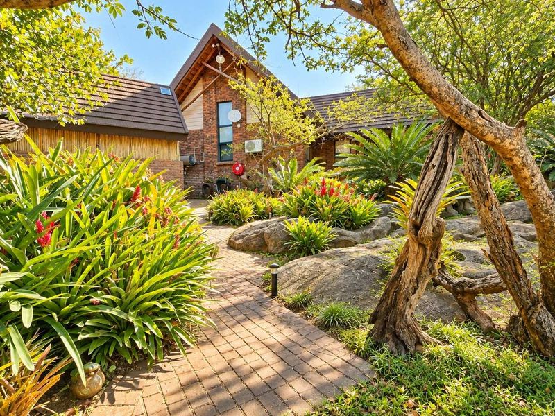 A charming brick-faced home entrance featuring a paved walkway, established landscaped gardens with natural rock features, and a pitched tiled roof.