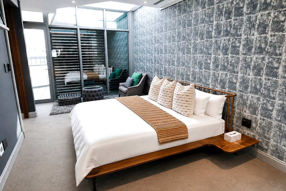 Modern penthouse bedroom featuring textured feature wall, recessed lighting, ducted air conditioning, and large glass sliding doors leading to a private balcony.