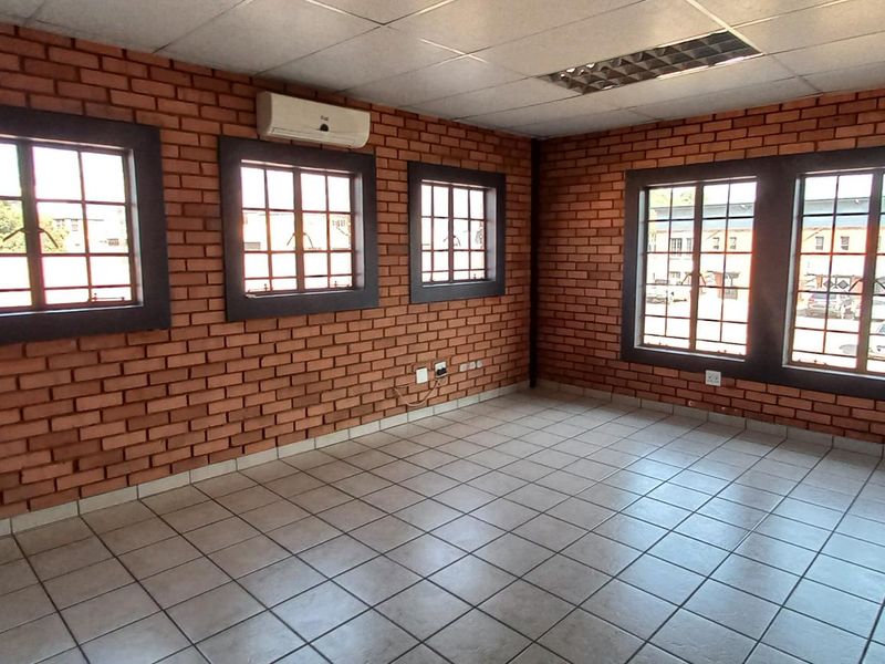 A spacious industrial office area featuring exposed brick walls, tiled flooring, multiple windows, and a wall-mounted air conditioning unit.