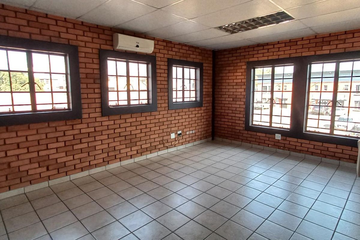 A spacious industrial office area featuring exposed brick walls, tiled flooring, multiple windows, and a wall-mounted air conditioning unit.