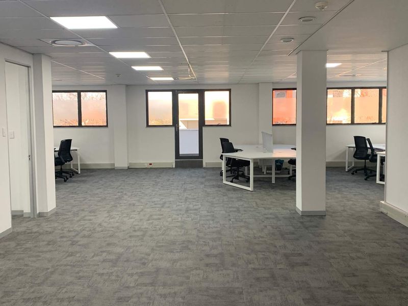 A spacious, well-lit open-plan office area featuring a suspended ceiling with recessed lighting, carpeted flooring, and large windows providing natural light.