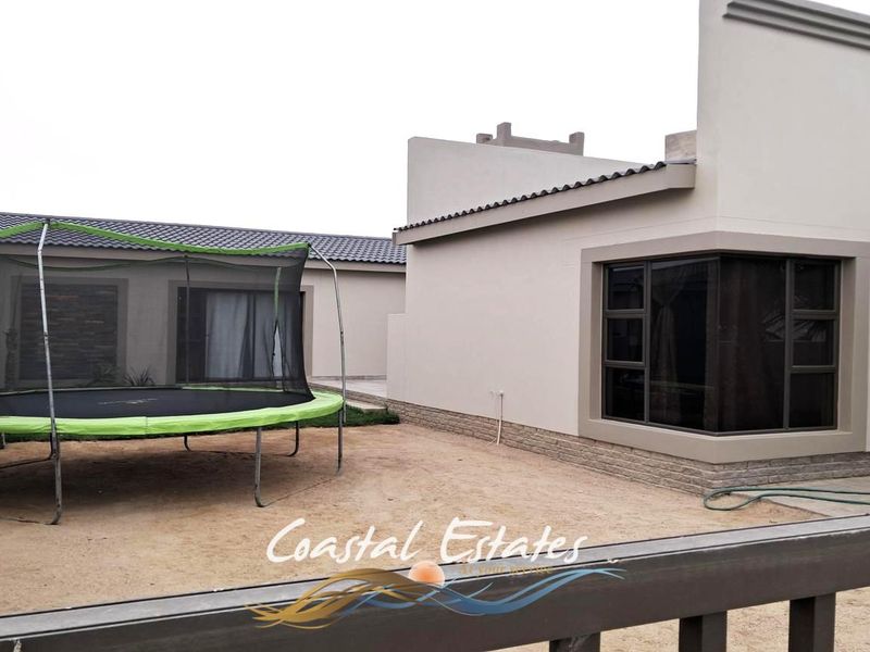 Rear view of the property featuring plastered walls, stone cladding accents, and dark aluminium window frames with a paved and sandy outdoor area.