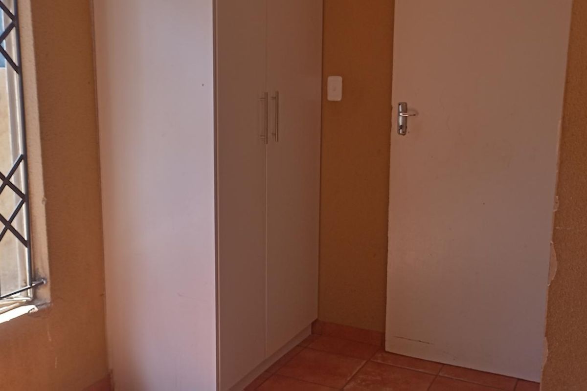 A bedroom featuring built-in cupboards, tiled flooring, and a window secured with metal burglar bars.