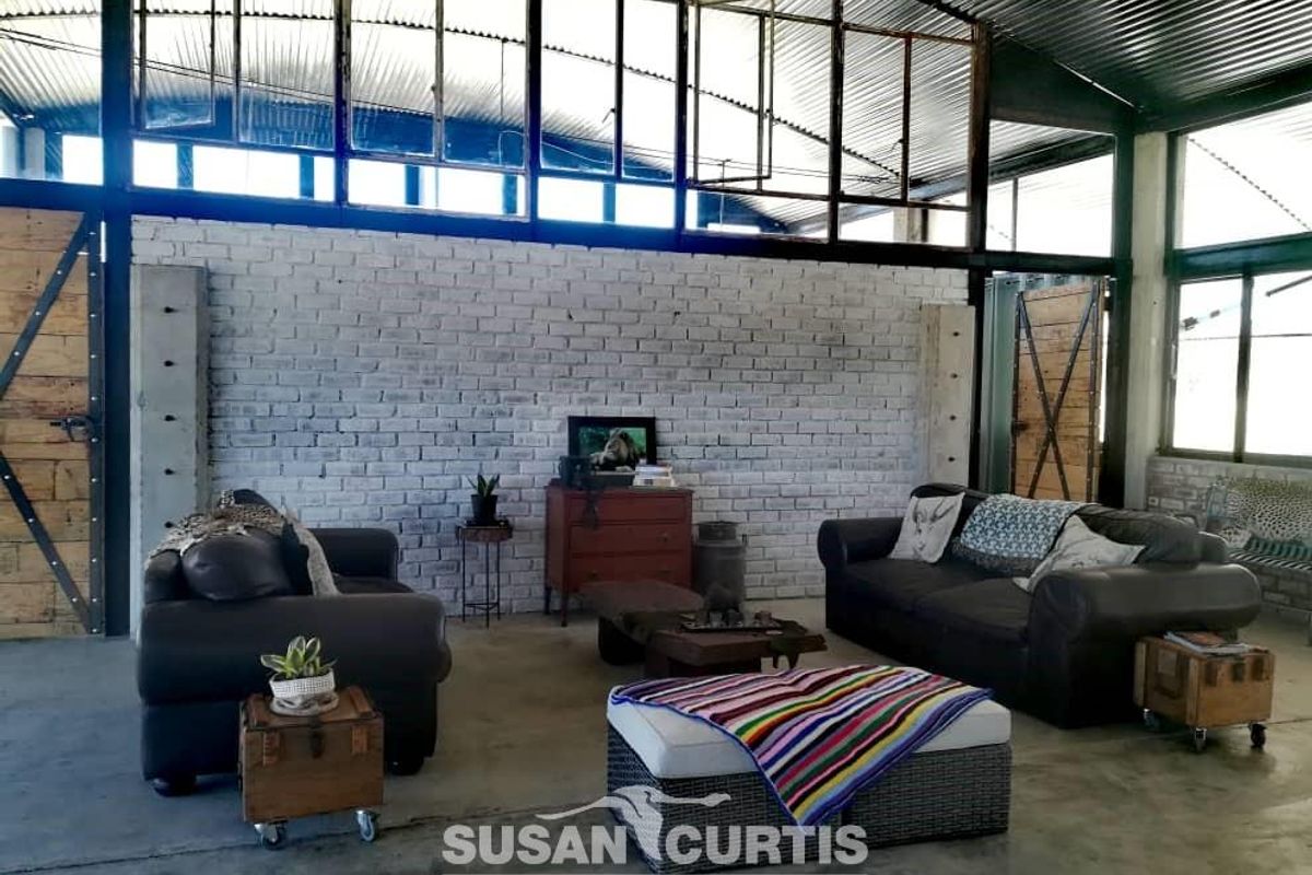 Spacious industrial-style living area featuring exposed brick walls, high vaulted corrugated metal ceiling, and large steel-framed windows providing ample natural light.