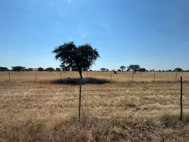 A wide-angle view of open farm land featuring dry grass, scattered trees, and perimeter wire fencing.