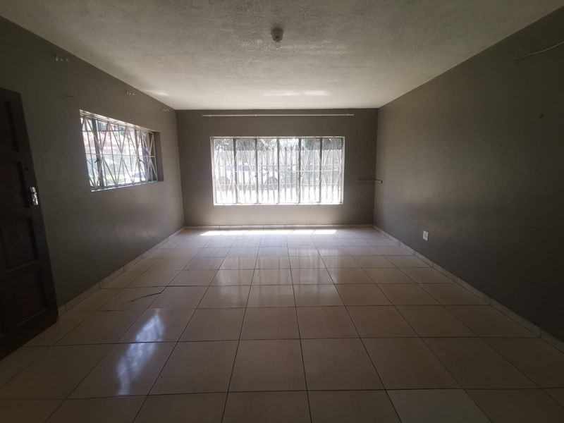 A spacious, empty living area featuring neutral tiled flooring, large windows with security bars, and dark-toned walls, offering a blank canvas for interior design.