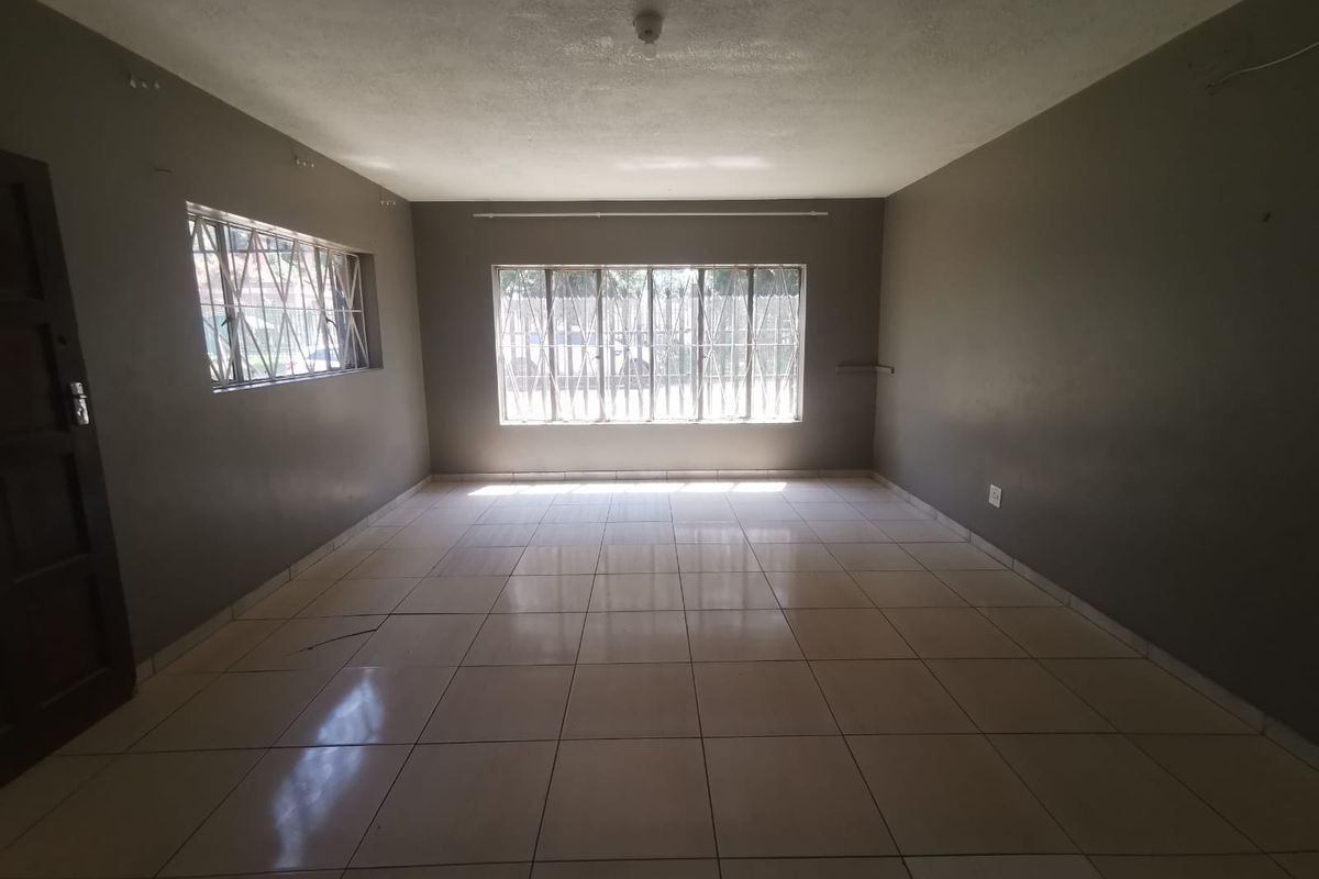 A spacious, empty living area featuring neutral tiled flooring, large windows with security bars, and dark-toned walls, offering a blank canvas for interior design.