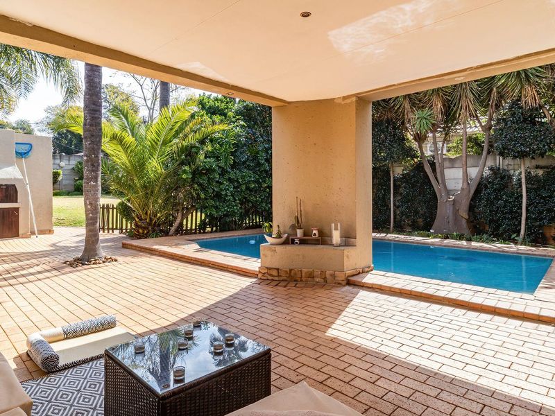Covered outdoor patio featuring brick paving, a built-in braai area, and direct access to a private swimming pool in a landscaped garden setting.